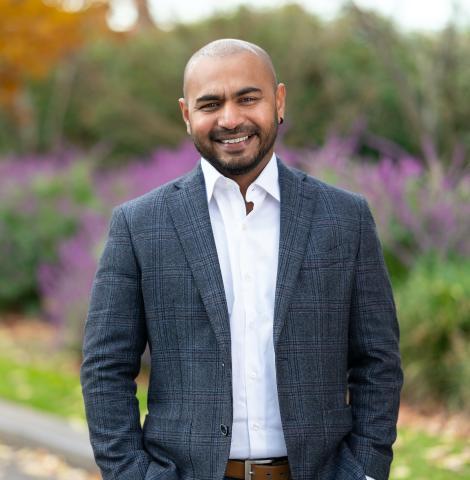 A man with a shaved head and trimmed beard, wearing a grey blazer and white button up shirt, Smiling in a park.